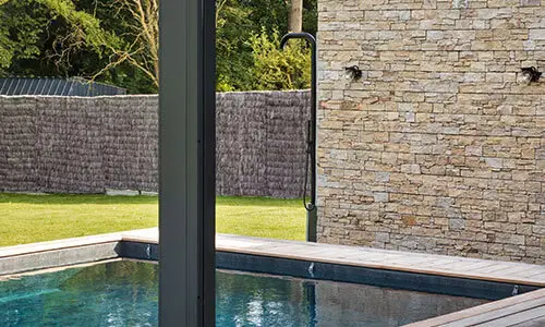 Piscine contemporaine entourée d’une terrasse en bois, avec mur en pierre naturelle et douche extérieure dans un cadre verdoyant.