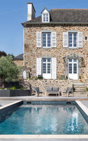 Piscine contemporaine construite par Nymfeo devant une maison en pierre traditionnelle avec volets blancs.