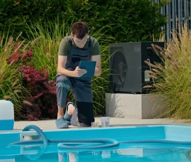 Technicien de piscine en tenue de travail accroupi au bord d’une piscine, utilisant une tablette pour analyser l’eau à côté d’un kit de test et de produits chimiques, avec une pompe à chaleur visible à l’arrière et un décor paysager verdoyant.